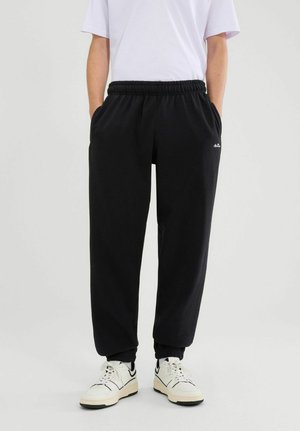 Osoba ubrana w czarne spodnie dresowe z elastycznym pasem i kieszeniami oraz białe sneakersy, stojąca na tle jasnego, jednolitego tła.