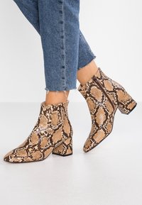 Botines de tobillo con estampado de serpiente, un patrón texturizado en beige y marrón, punteras afiladas y un tacón cuadrado, combinados con unos jeans de mezclilla cortos.