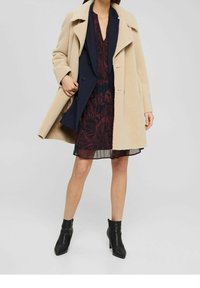 Beige ullblandad kappa med breda slag, lagd över en marinblå jacka och en vinröd mönstrad klänning. Svarta ankelskor med spetsig tå.