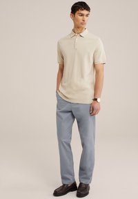 Bež majica s kratkimi rokavi, s tremi gumbi na zapenjanju, v kombinaciji z svetlo modrimi hlačami in temno rjavimi moškimi čevlji. Gladka površina tkanine.