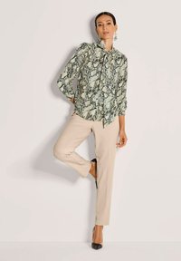 Frau in einer Schlangenlederbluse, beigen Hosen und schwarzen Absatzschuhen, die gegen eine helle Wand steht, mit einem gebeugten Bein und einer Hand auf der Hüfte.