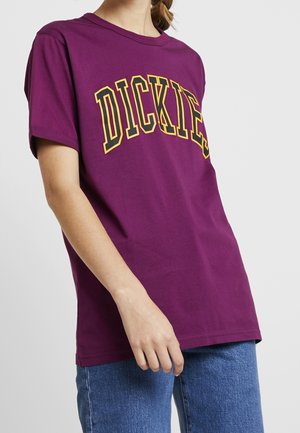 Lilla bomulds T-shirt med fed gul og sort "DICKIES" tekst hen over brystet, korte ærmer og en afslappet pasform.