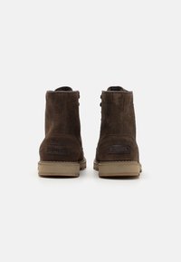 Botas de tobillo de ante marrón con suelas de goma plana color yute. Presentan un diseño cosido y una etiqueta de marca en la parte trasera. Tienen dos tiradores en la parte superior.