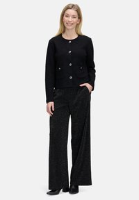 Kobieta ubrana w czarną, teksturowaną kurtkę na guziki, błyszczące szerokie czarne spodnie oraz czarne botki na obcasie, uśmiechająca się z jedną ręką w kieszeni.