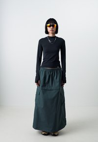 Haut à manches longues noir associé à une jupe maxi verte avec taille élastique, détails de poches et texture lisse. Le modèle porte des lunettes de soleil.