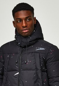 Μαύρο μπουφάν puffer με ψηλό γιακά, με πιτσιλιές λευκού, ροζ και μπλε χρώματος. Μπροστινό φερμουάρ και ρυθμιζόμενη κουκούλα με κορδόνι.