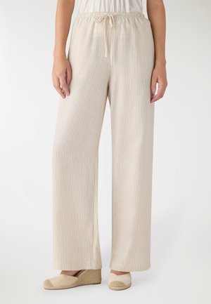 Donna che indossa pantaloni beige a gamba larga con coulisse in vita e scarpe espadrillas beige con zeppa, in piedi su sfondo bianco.