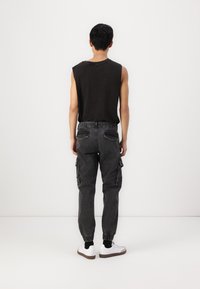 Zwarte mouwloze shirt, donkergrijze cargobroek met elastische boorden, zijzakken en witte sneakers met zwarte accenten, gezien van achteren.