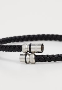 Bracelet en cuir tressé noir avec des accents en métal argenté, présentant un matériel texturé et une marque gravée. Deux extrémités de fermoir sont visibles.