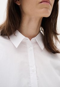 Kobieta w białej koszuli zapinanej na guziki z kołnierzykiem, ukazująca swoją szyję, podbródek i częściowo usta, na jednolitym jasnym tle.
