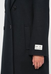 Abrigo de lana negro con un diseño de doble botonadura, botones grandes y un bolsillo lateral. Una etiqueta con texto es visible en la manga.