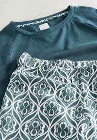 T-Shirt aus Baumwolle in Petrol mit Rundhalsausschnitt, kombiniert mit grünen Shorts mit Muster, die weiße geometrische Designs und einen elastischen Bund haben.