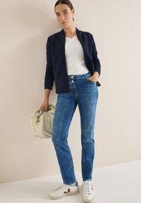 Námořnicky modrá bomber bunda přes bílé tričko, modré džíny a bílé tenisky s červenými detaily. Model drží světle zelenou tašku.