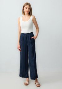 Witte geribde tanktop gecombineerd met marineblauwe brede broek met zijzakken, van gladde stof, en zachte beige instapsandalen.