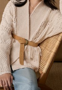 Béžový pletený cardigan s texturovanými vzory, zpevněný hnědým koženým páskem. Nosit na světle modrých džínech, sedící na tkané židli.