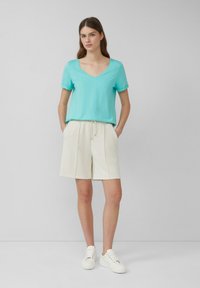 Turkusowa koszulka typu V-neck z podwiniętymi rękawami, w połączeniu z beżowymi szortami na sznurku. Model nosi białe sneakersy, stoi na tle w neutralnych kolorach.