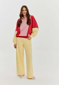 Barevný svetr s dlouhým rukávem v červené, růžové a žluté barvě, doplněný žlutými širokými kalhotami. Tkanina vypadá jemně a pohodlně, s volným střihem.