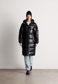 Garš melns pūšamais mētelis ar spīdīgu apdari, pārmērīgs izmērs, lielas kabatas un kapuce, apvienots ar gaišiem ziliem džinsiem un melnām klompām.