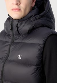Zwarte puffer vest met capuchon, voorzien van een ritssluiting aan de voorkant, matte afwerking, gewatteerde textuur en een witte "CK"-logo op de borst.