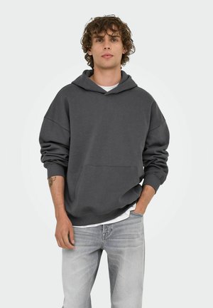 Young man with curly hair wearing a dark gray hoodie over a white shirt and light gray jeans, standing with one hand in pocket.