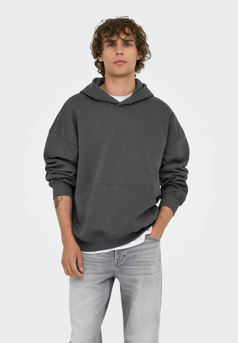 Joven con pelo rizado que lleva una sudadera gris oscuro sobre una camisa blanca y jeans gris claro, de pie con una mano en el bolsillo.
