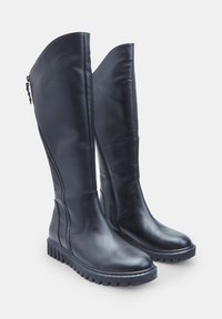 Botas largas de cuero negro con cremalleras laterales y suelas de goma gruesa y estriada, de pie sobre un fondo blanco.