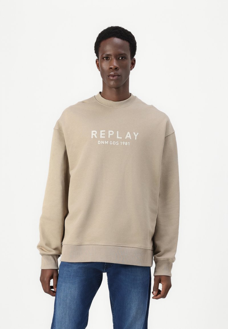 Beige pulóver kerek nyakkivágással és hosszú ujjakkal. Elején fehér nyomtatott szöveg "REPLAY DNM GDS 1981" található. Rugalmas mandzsetták és alsó szegély.