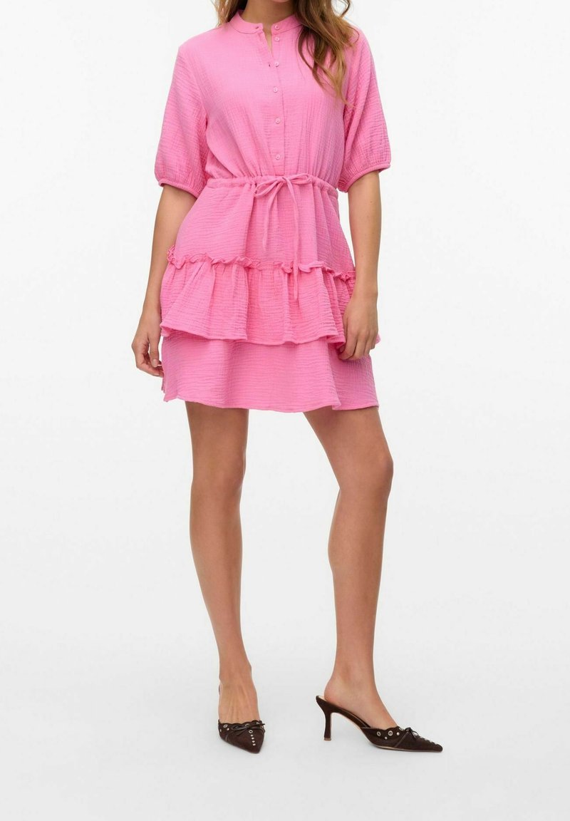 Femme portant une robe rose texturée avec des manches bouffantes, un devant boutonné, une ceinture à la taille, un ourlet à volants, et des mules à talons noires à bout pointu.