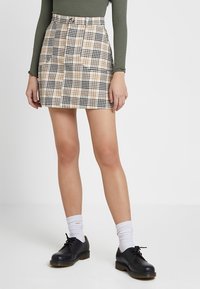 Minifalda de cuadros en beige, negro y blanco, con una cintura alta y bolsillos visibles. Llevada con zapatos negros y calcetines blancos.