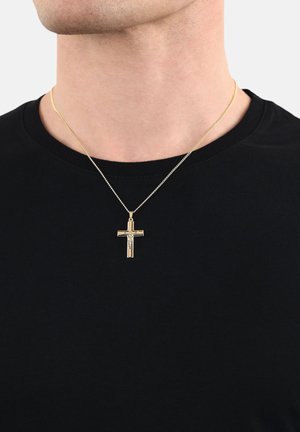Collier avec pendentif croix en ton doré sur une fine chaîne, porté sur un t-shirt noir. La croix présente des détails complexes avec une finition texturée.