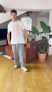 Biely grafický tričko s ružovým potlačou, sivé tepláky a biele tenisky. Prostredie obsahuje hnedú kordurovú pohovku a zelenú rastlinu.