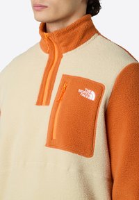 Fleece pullover in crème en oranje, met een hoge kraag, een ritszak op de borst en een logodetail. Zachte textuur met contrasterende kleuren.