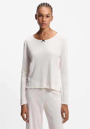 Vrouw draagt een witte getextureerde lange-mouwen top met zwarte strikdetail en bijpassende broek, staand tegen een effen lichte achtergrond.