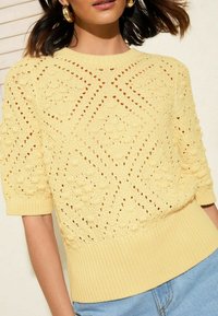 Suéter de punto amarillo claro con un patrón texturizado en forma de diamante, puños y dobladillo acanalados. Presenta mangas cortas y un diseño transpirable.