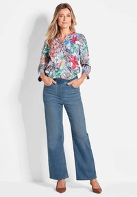 Blumenbluse mit bunten Mustern, langen Ärmeln und V-Ausschnitt; kombiniert mit blauen weit geschnittenen Jeans und braunen Ballerinas.