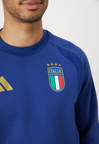Plava dukserica s okruglim izrezom, koja sadrži zlatni Adidas logo i grb talijanske nogometne reprezentacije s četiri zvjezdice. Teksturalni akcenti na ramenima.
