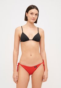 Crni trokutasti bikini gornji dio s tankim trakama u paru s crvenim bikini donjim dijelom koji ima bočne vezice i mali logotip s prednje strane.