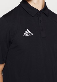 Czarna koszulka polo z gładkiego materiału z kołnierzykiem, posiadająca trzy guziki oraz białe haftowane logo Adidas po lewej stronie.