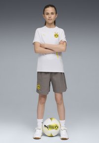 Vit sportskjorta med gula detaljer, grå shorts med en logotyp, vita och svarta sneakers samt en mönstrad fotboll.