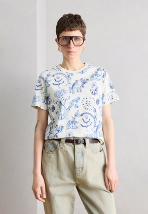 Personne aux cheveux courts bruns et grandes lunettes porte une chemise blanche à manches courtes avec des imprimés bleus floraux et animaux, un pantalon beige et des boucles d'oreilles en or.