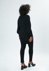Schwarzes, geripptes Strickkleid mit langen Ärmeln, figurbetontem Schnitt und mittellanger Länge bis zur Oberschenkelmitte, kombiniert mit schwarzen, eng anliegenden Hosen und klobigen, hohen Sandalen.
