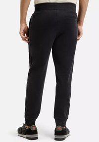 Mężczyzna w czarnych dresowych spodniach i czarnych sneakersach, stojący z rękami luźno wzdłuż ciała, zwrócony tyłem do aparatu na białym tle.