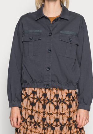 Kvinde iført en mørk knappet jakke med brystlommer og elastiske manchetter over et mønstret orange og sort skørt.