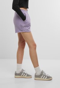 Pantaloni corti di cotone viola con orlo sfrangiato, dotati di tasche laterali e dettaglio ricamato a farfalla. Indossati con sneakers grigie e calzini bianchi.