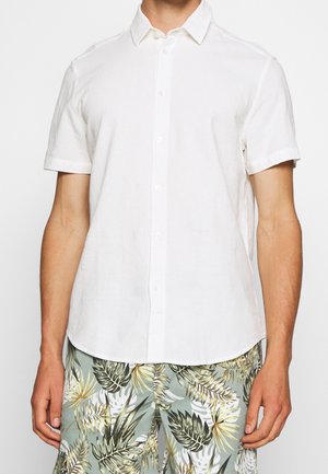 Homme portant une chemise blanche unie à manches courtes et un short vert clair à imprimé tropical avec des motifs de feuilles.