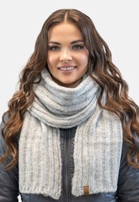 Écharpe en tricot gris clair avec une texture côtelée, présentant une finition douce et une petite étiquette en cuir. Portée sur une doudoune noire.