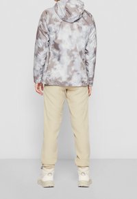 Grijze en witte camouflage-jack met een capuchon, gecombineerd met beige broek en lichte schoenen. Soepele, reflecterende stoftextuur.