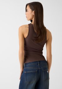 Bruine ribgebreide tanktop gecombineerd met donkerblauwe spijkerbroek met achterzakken en een logopatch. Het model staat met de rug naar voren, waardoor de pasvorm van de outfit duidelijk zichtbaar is.