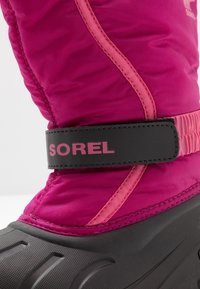 Botte isolante rose avec semelle en caoutchouc noir texturé et sangle noire portant "SOREL" en rose. Coutures latérales en rose contrastant.