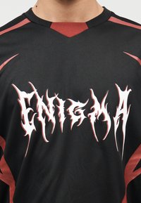 Μαύρο δίχτυ αθλητική φανέλα με v-λαιμόκοψη σε κόκκινη απόχρωση. Μπροστά εμφανίζεται η λέξη «ENIGMA» σε λευκή, αιχμηρή γραμματοσειρά.